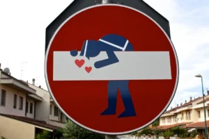Immagine che mostra un cartello stradale di divieto d'accesso modificato con un adesivo che raffigura una sagoma stilizzata intenta a baciare la barra bianca centrale, circondata da tre cuori rossi, usata simbolicamente per riferirsi al modo d'intendere la dipendenza affettiva proposto da Alessio Congiu come un blocco nello sviluppo del rapporto sentimentale