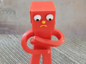 Foto di un pupazzo di plastica rosso con testa cubica, occhi mobili, naso giallo e bocca triste, ritratto a braccia conserte, usato per riferirsi simbolicamente ai disagi psico-fisici dell'astinenza affettiva, tema di cui si occupa a Cagliari, Verona e online il Dr. Alessio Congiu